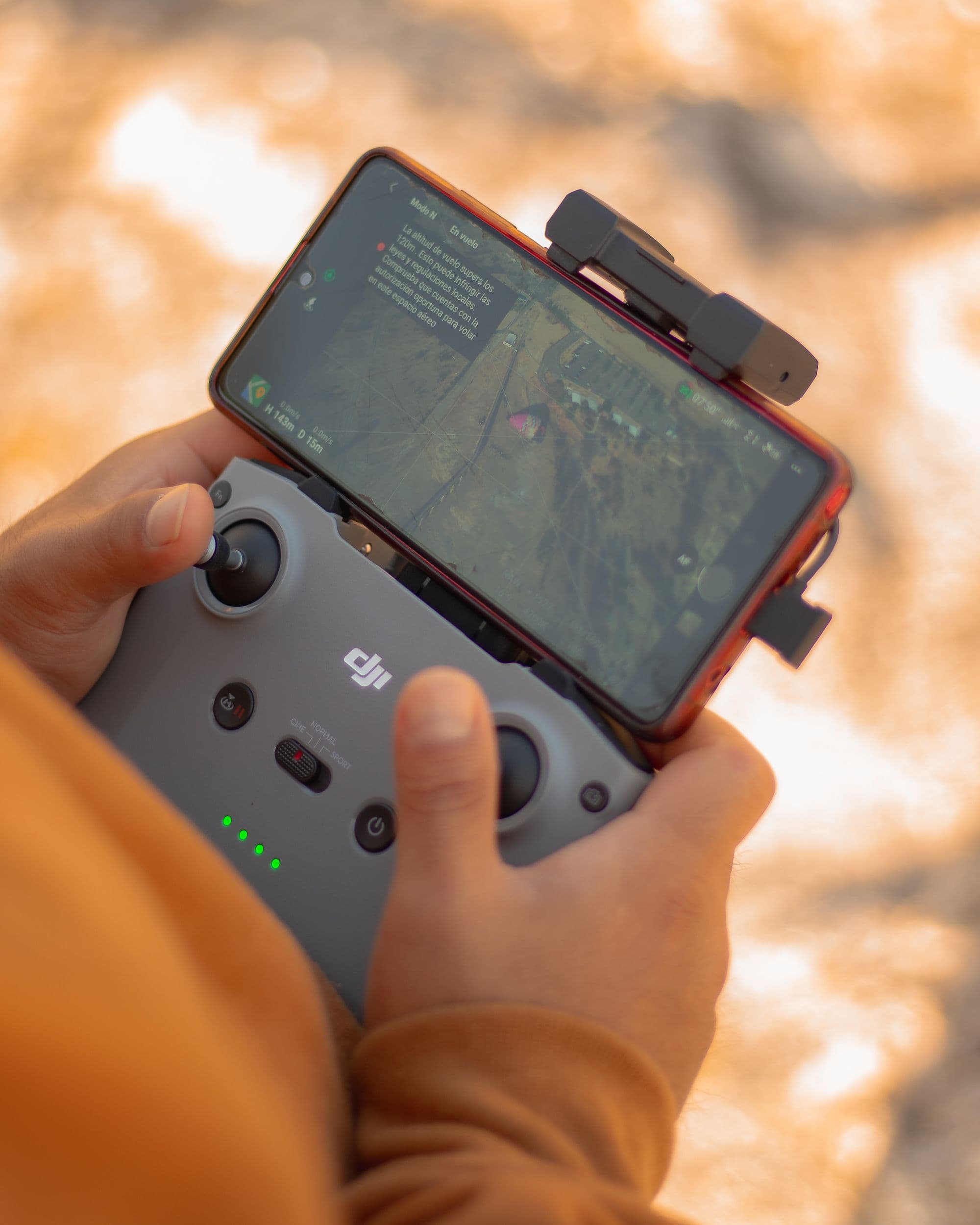 A pilot holding a drone controller and phone outdoors in warm evening light.